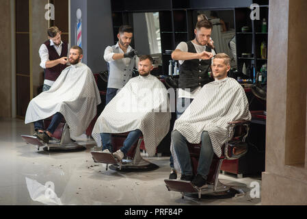 Processo di styling e della cura del corpo maschile di parrucchieri in barberia. Tre professionale e fiduciosi i barbieri in piedi e il taglio di capelli di uomini. Dei clienti di sesso maschile seduto in sedie e taglio di capelli da indossare gli abiti. Foto Stock