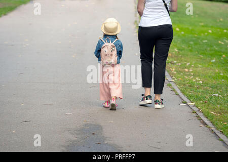 Bambina con uno zaino e la mamma andare sul marciapiede. Vista dal retro. Foto Stock