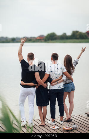 Compagnia di giovani in piedi, guardando il lago, vista posteriore. Amici godere di riposo nella natura, sul molo di legno in fiume Foto Stock