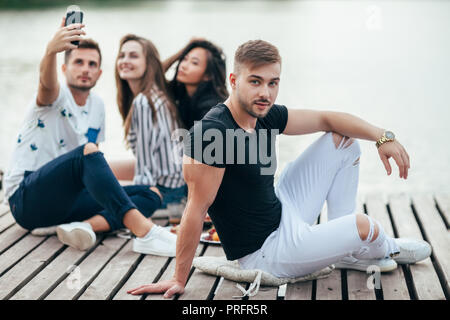 Bel giovane rilassante sul molo in legno. I suoi amici in background facendo selfie. Vacanze, tempo libero e spensierato passatempo concept Foto Stock