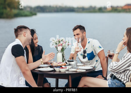Gruppo di felice giovani amici di mangiare e divertirsi in all'aperto ristorante Riverside. Cibo, divertimento e concetto di pranzo Foto Stock