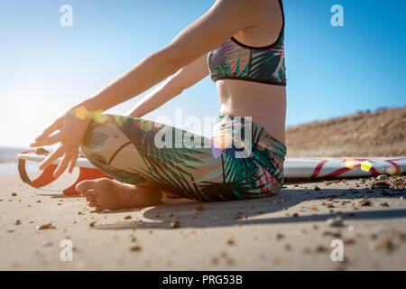 Surfista femmina seduto sulla spiaggia meditando la pratica dello yoga prima di iniziare la navigazione Foto Stock