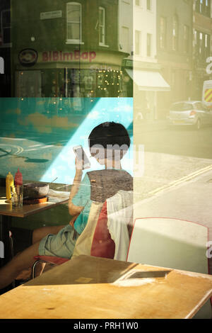 Attraverso la finestra - ragazza asiatica guardando il suo cellulare a tavola da pranzo,Soho,Londra Foto Stock