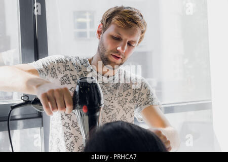 Professional sorridente stilista maschio soffiare l'asciugatura donna capelli con un essiccatore in salone Foto Stock