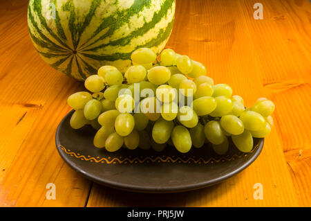 Still Life - un pennello grande di uva verde e anguria in un buio piastra ceramica su uno sfondo di legno Foto Stock