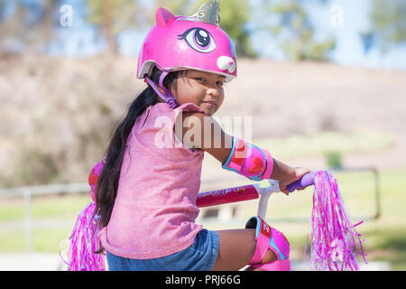 5 anno vecchia ragazza ottenere pronto per cavalcare lontano nel suo moto rosa, indossando il casco protettivo e ginocchiere in un parco con le colline Foto Stock