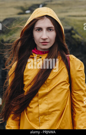 Attraente giovane donna in impermeabile giallo guardando la telecamera con le montagne sullo sfondo Foto Stock