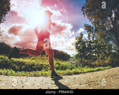 Jogging di altezza uomo sportivo in alberi ombre con luce del sole dietro di lui mentre vestita di nero pantaloncini giallo e blu abbigliamento per fare jogging Foto Stock