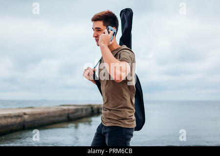 Giovane uomo che porta la chitarra acustica in casw e parlando al telefono camminando sulla spiaggia nuvoloso Foto Stock