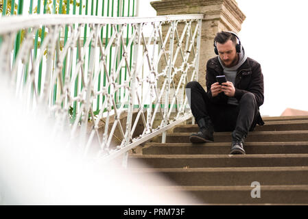Un bel giovane hipster uomo seduto sulle scale e controllando il suo smartphone. Foto Stock