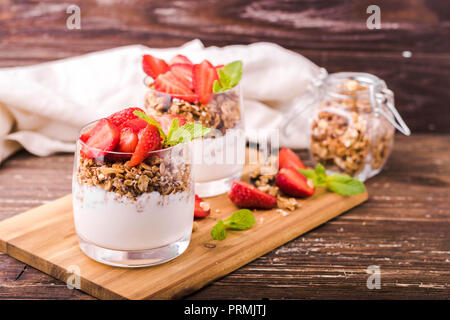 Parfait di yogurt in bicchieri con muesli, fragole e foglie di menta. Sulla tavola di legno. Colazione sana nozione. Tavola in legno rustico sfondo. Foto Stock