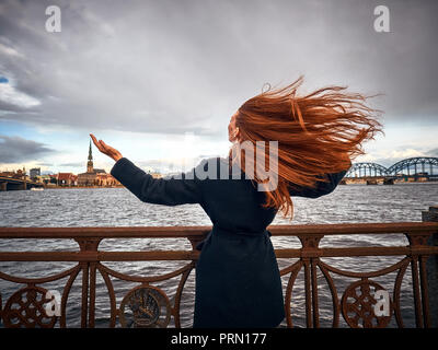 Una giovane ragazza rivolta verso la città ha sollevato le sue mani verso l'alto. I suoi lunghi capelli rossi si sviluppa nel vento. La ragazza è stata fotografata dal retro. Foto Stock