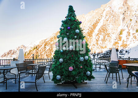 Bellissimo albero di Natale decorato con le palline e ghirlande sullo sfondo delle montagne in un cafe' all'aperto con la luce naturale in alta montagna Foto Stock