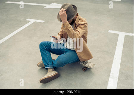 Angolo di alta vista di elegante uomo in hat seduta su skateboard e utilizza lo smartphone Foto Stock