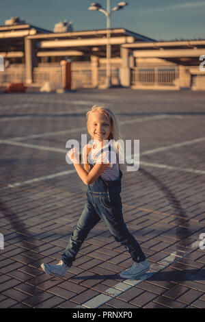 Sorridente bambino adorabile camminando con lo skateboard in mani e guardando la telecamera presso il parcheggio Foto Stock