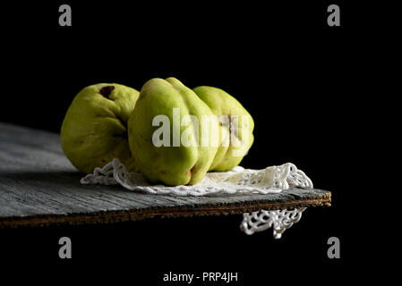 Primo piano di alcuni frutti di mela cotogna, su un centrino a crochet, posto sul grigio di una tavola in legno rustico, contro uno sfondo nero Foto Stock