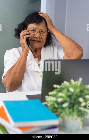 Ha sottolineato il senior donna parlando al telefono cellulare e di lavoro sul computer portatile Foto Stock