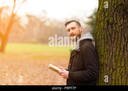 Un sorridente bel giovane hipster uomo appoggiato a un albero e la lettura di un libro nel parco. Foto Stock