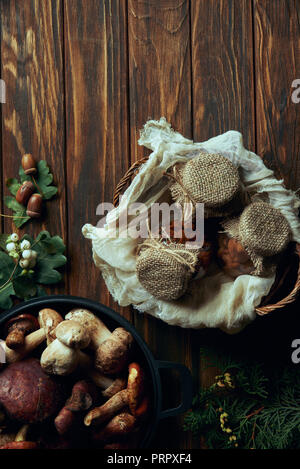 Vista dall'alto di deliziosi funghi sott'olio nel cestello e di funghi freschi di coppa sul tavolo di legno Foto Stock