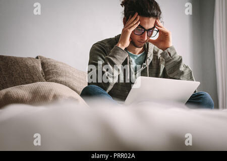 Uomo seduto a casa in teso umore guardando il laptop. Uomo che guarda preoccupato seduta sul letto con mani toccano la testa. Foto Stock