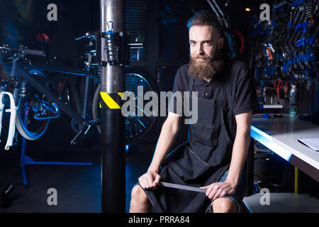 Ritratto di proprietario di piccola impresa del giovane uomo con la barba. Guy bicicletta officina meccanica lavoratore seduto con lo strumento in mano a un gruppo di lavoro di vestire nero Foto Stock