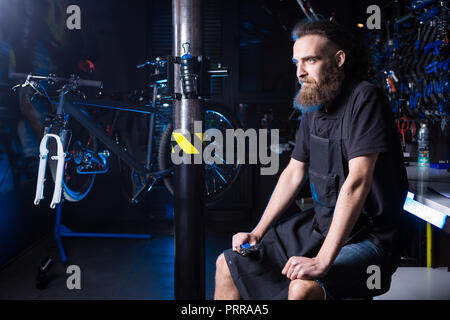 Ritratto di proprietario di piccola impresa del giovane uomo con la barba. Guy bicicletta officina meccanica lavoratore seduto con lo strumento in mano a un gruppo di lavoro di vestire nero Foto Stock