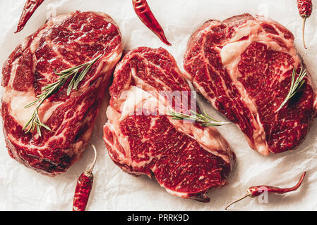 Vista superiore di tre pezzi di carne cruda bistecche, peperoncino e rosmarino sulla carta da forno in cucina Foto Stock