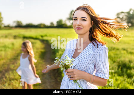 Giovane e bella madre con fiori di campo bouquet e piccola figlia sfocati sullo sfondo nel campo verde Foto Stock