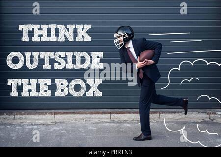Vista laterale di imprenditore in tuta e casco di rugby con la palla in mano in esecuzione su strada con think outside box iscrizione Foto Stock