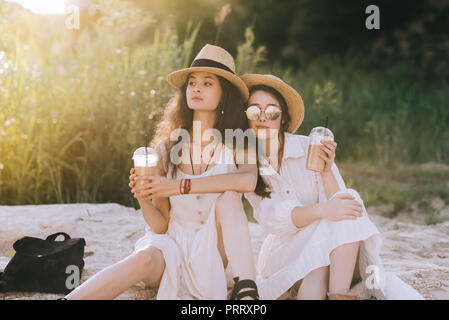 Bellissima femmina amici in cappelli di paglia tenendo i bicchieri di plastica con caffè latte e sedersi sul terreno con la luce solare Foto Stock