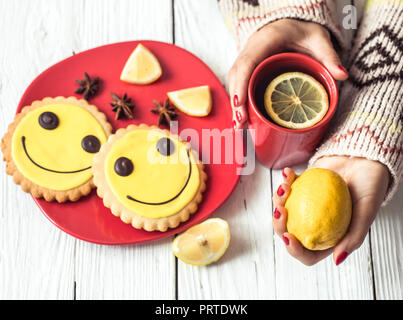 Red tazza di tè caldo con limone nelle mani della ragazza in il maglione,su un bianco sullo sfondo di legno rosso e una piastra di cookie,il concetto di bevande calde Foto Stock