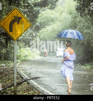 Una giovane ragazza in un abito sotto la pioggia con un ombrello lungo la strada Foto Stock