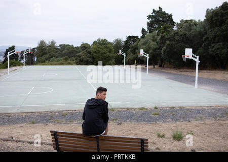 Un adolescente di sesso maschile del giocatore di basket guardando indietro da una panchina nel parco dal campo di pallacanestro Foto Stock
