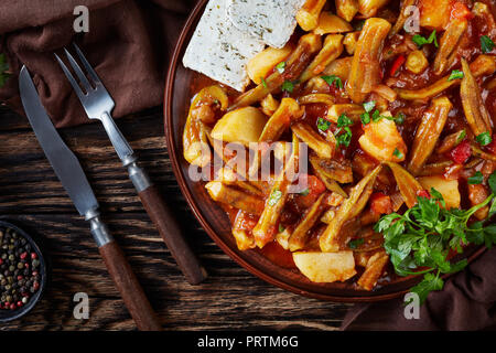 L'okra stufato di pomodoro con patate servita con il formaggio feta le fette su un piatto di terracotta su un tavolo rustico, ricetta greca. forcella, coltello, prezzemolo in backgr Foto Stock