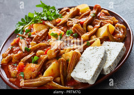 L'okra stufati con pomodori e patate servita con il formaggio feta le fette su un piatto di terracotta su un calcestruzzo di colore grigio nella tabella, ricetta greca, vista da sopra, Foto Stock