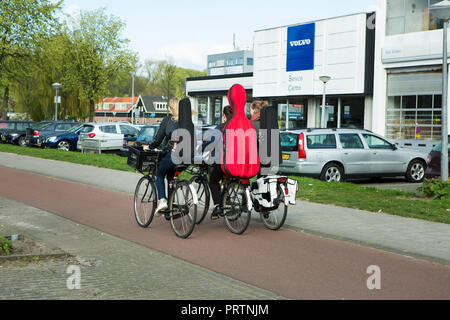 Tre i ciclisti che trasportano il violino e violoncello casi pedalando attraverso un concessionario Volvo segno. Amsterdam. I Paesi Bassi. Foto Stock