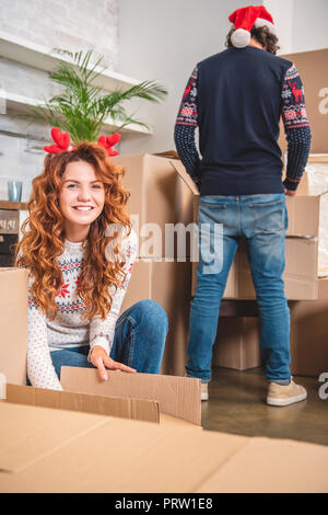 Felice giovane donna in palchi con archetto disimballaggio di scatole di cartone e sorridente alla fotocamera mentre il marito in santa hat in piedi dietro Foto Stock