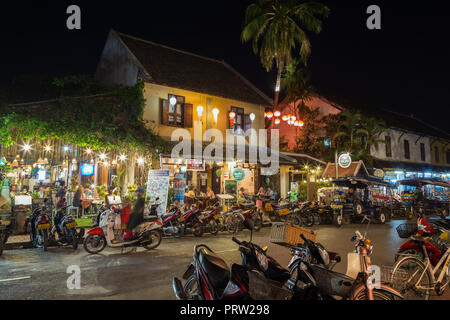 Scooter a tre ruote taxi (jumbo o tuk-tuk) e turisti in caffetterie idilliaco in francese era coloniale edifici a Luang Prabang, Laos, di notte. Foto Stock