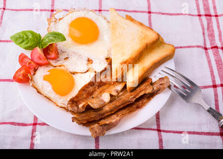 La prima colazione con pancetta e uova fritte. Servita su piastra bianca con fette di pomodoro ciliegino, foglie di basilico e toast croccanti slice.