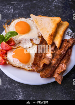 La prima colazione con pancetta e uova fritte. Servita su piastra bianca con fette di pomodoro ciliegino, foglie di basilico e toast croccanti slice. Dark grunge tabella di calcestruzzo Foto Stock