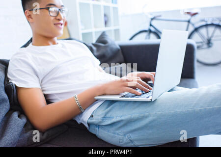 Vista laterale di allegro bello uomo asiatico utilizzando computer portatile sul divano di casa Foto Stock