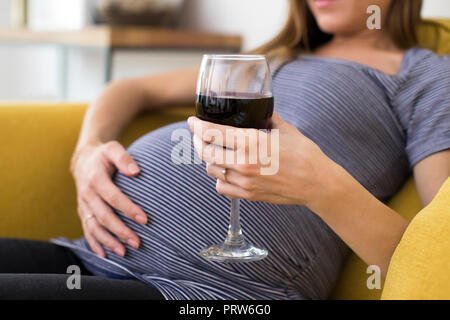 Close up della donna incinta di bere vino rosso seduto sul divano di casa Foto Stock