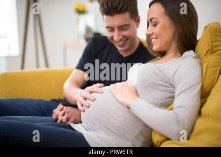 Incinta giovane seduto sul divano con uomo toccando Womans Stomaco Foto Stock