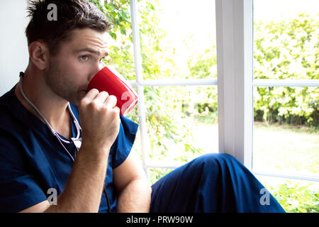 Bel maschio medico o infermiere in pausa indossando blue frega di bere dalla tazza e guardando fuori della finestra Foto Stock