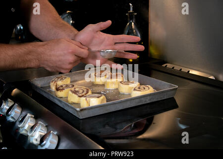 Muscolo forti braccia dei maschi di chef, baker polvere di cannella sulle ciambelle alla cannella, turbolenze hot fuori del forno Foto Stock