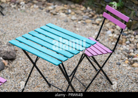 Una rosa di legno sedia pieghevole e un quadrato tavolo blu sono in piedi sulla ghiaia nel cortile di una caffetteria nella luce naturale di sera Foto Stock