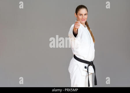 Fiducioso athletic giovane donna in kimono bianco e nero permanente della cinghia, il puntare il dito e guardando la fotocamera con sorriso. Giapponese di arti marziali concep Foto Stock