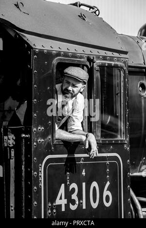 Ritratto, bianco e nero primo piano del macchinista, all'interno della cabina della locomotiva a vapore britannica d'epoca inclinata e con vista sul finestrino del motore in estate sole. Foto Stock