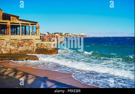 A piedi lungo la spiaggia di sabbia di Dahab resort con una vista sulla fascia costiera ristoranti, alberghi e fumoso montagne di Saudy Arabia, visto dietro il mare, il peccato Foto Stock