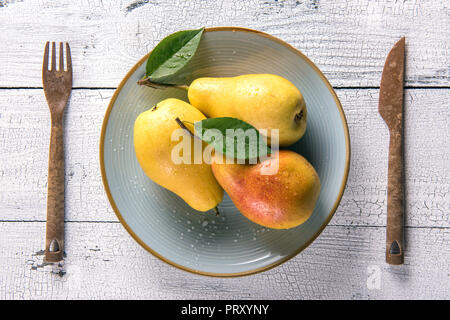 Umido giallo pere con foglie sul round piastra blu e sul vecchio incrinato bianco tavolo in legno con coltello e forchetta su entrambi i lati. Messa a fuoco selettiva. Vista da una Foto Stock